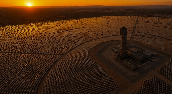 Vista aérea de una planta termosolar de torre central rodeada por miles de heliostatos, bajo un cielo anaranjado al atardecer.